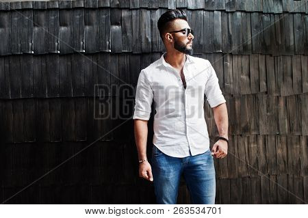 Stylish Tall Arabian Man Model In White Shirt, Jeans And Sunglasses Posed Against Wooden Wall Indoor