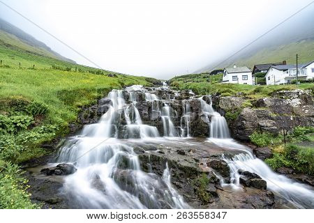 Beautiful And Peaceful Waterfall (long Exposure) With Green Grass And Small Houses In Foggy Weather 