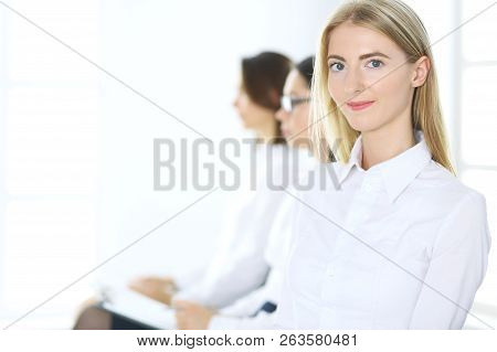 Attractive Businesswoman At Meeting Or Conference Against The Background Of Colleagues. Group Of Bus
