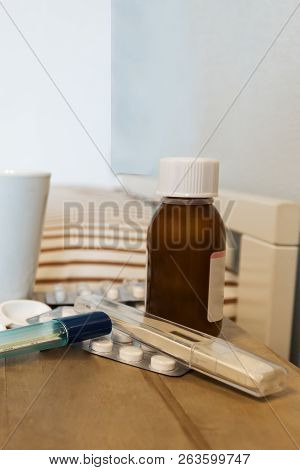 Close Up View Over A Wooden Table Near Bed With Syrip, Pills, Capsules And Thermometer On It. Flu An