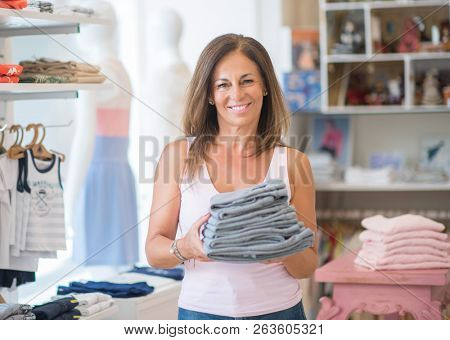 Beautiful middle age hispanic woman standing with smile on face at clothes store. Shop owner and shop assistant smiling confident and cheerful holding clothes.