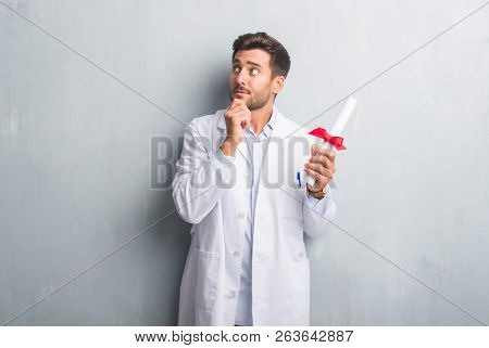Handsome young doctor man over grey grunge wall holding diploma serious face thinking about question, very confused idea