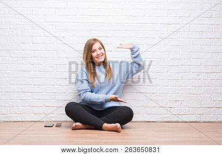 Young adult woman sitting on the floor in autumn over white brick wall gesturing with hands showing big and large size sign, measure symbol. Smiling looking at the camera. Measuring concept.