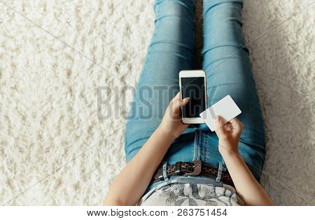 Young Woman Holding Credit Card And Using Phone Sitting On Floor. Payment.  Online Shopping Concept.