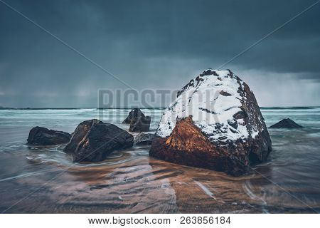 Rocks covered with snow on Norwegian sea beach in fjord in stormy weather with clouds. Skagsanden beach, Flakstad, Lofoten islands, Norway. Long exposure motion blur