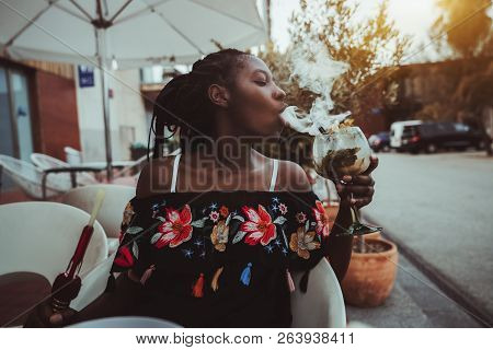Portrait Of A Charming African Girl Sitting In A Street Cafe And Making Smoke In The Glass Of Cockta