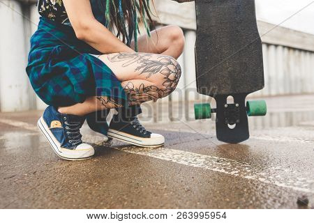 Young Girl With Tattoos And Dreadlocks Sits With A Longboard On The Background Of Concrete Structure