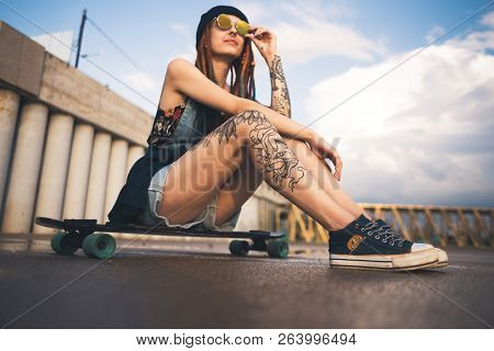 Young Girl With Tattoos And Dreadlocks In A Blue Cap Sits On A Longboard Against The Background Of C