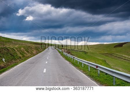 Asphalt Road Leading To The Top Of The Hill, Vibrant Green Meadow At Springtime With Dramatic Stormc