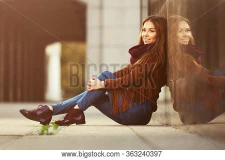 Happy young fashion woman sitting on street sidewalk  Stylish female model in brown leather fringe suede jacket and dark blue jeans