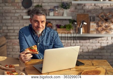 Man working from home on laptop computer, sitting at table in kitchen, eating online ordered pizza.