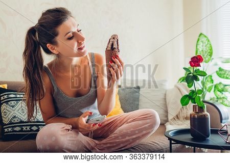 Young Woman Eating Chocolate Ice-cream In Cone Lying On Couch At Home And Using Smartphone. Relaxing