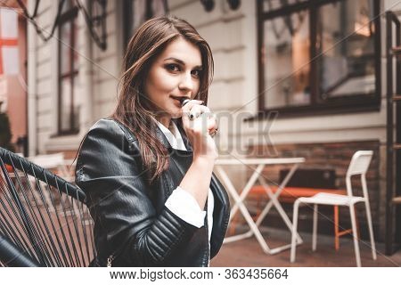 Positive Girl Sits On The Street At The Cafe Table And Smokes Electronic Cigarette.
