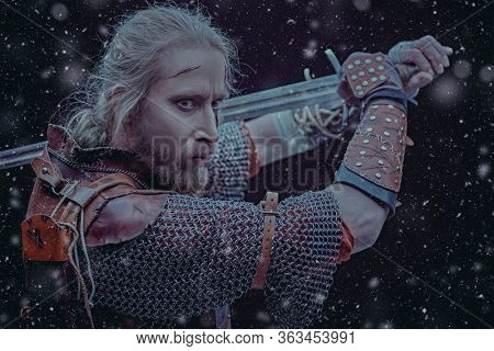 Portrait of a brave medieval warrior in armor with a sword on a black background with snowfall. The Witcher man.
