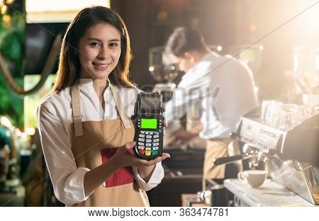 Asian Barista woman holding creadit card reader machine cashless payment using EMV chip technology for coffee purchase at cafe. Cashless preventing from coronavirus covid-19 Spreading and infection