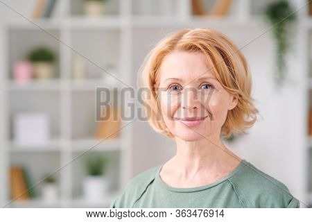 Smiling mature woman with under eye patches looking at you while standing in front of camera on background of shelves with flowerpots and books