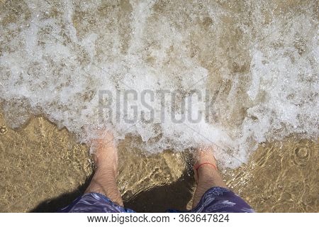 Top View Of Man's Feet (barefoot) On The Sand At The Surf Waves Over Them. Point Of View On Legs In 