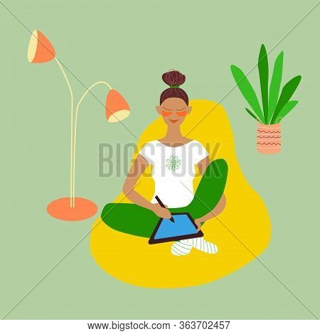 Girl Draws On A Tablet, Sitting In A Bean Bag; Floor Lamp And A Home Plant. Young Brunette Woman Wor