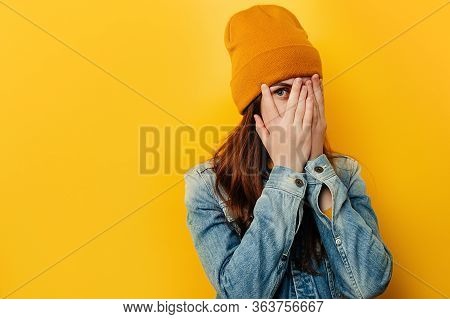 Shocked Frightened Young Woman Peeks Through Fingers, Stares With Widely Opened Eyes, Wears Denim Ja