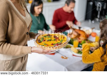 Woman Holding Thanksgiving Pie Near Blurred Family Celebrating Thanksgiving At Home.