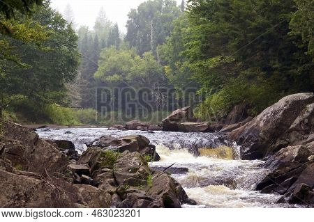 Beautiful Misty Waterfall Surrounded By Large Rocks And Boulders With Thick Dense Forest In Backgrou
