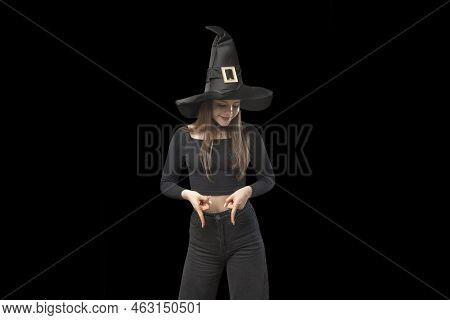 Girl In Pointed Hat Points Her Finger Down. Portrait Of Witch Girl In Isolation On Black Background.