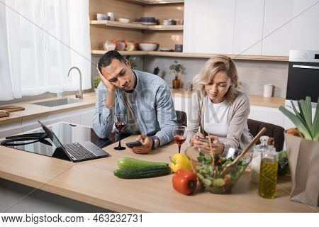 Tired, Emotionless Multinational Couple Eating Salad Using Smartphone Checking Photos On Social Medi
