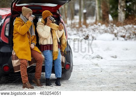 Full Length Portrait Of Young Couple Sitting In Car Trunk In Winter Forest While Enjoying Hot Cocoa,