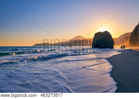 Silhouettes Of Two Adults Walking In Tide, Beach On Sunset, Milos, Greece. Iconic Fyriplaka Lonely R