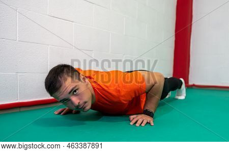 Determined Strong Young Man Doing Push-ups And Looking At Camera. He Is Dressed In An Orange Shirt A