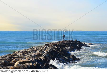 Fisherman On Masonry Breakwater. A Fisherman Catches Fish In The Sea On A Stone Breakwater. Waves At