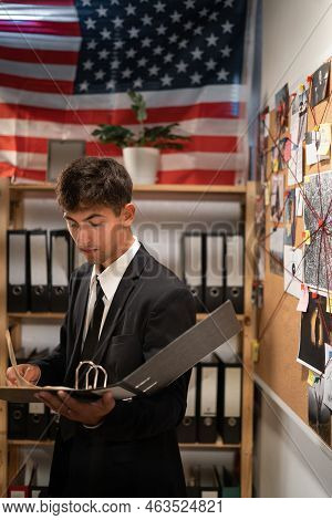 Concentrated Young Detective In Black Suit Reading Dossier In Dark Office. Investigator Working With