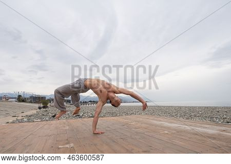 Shirtless Caucasian Man Doing Backflip On Pebble Beach.