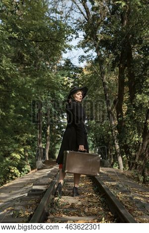 Woman In Black Coat, Hat And With Retro Suitcase Wallking On Rails In Forest. Vintage Clothing. Back