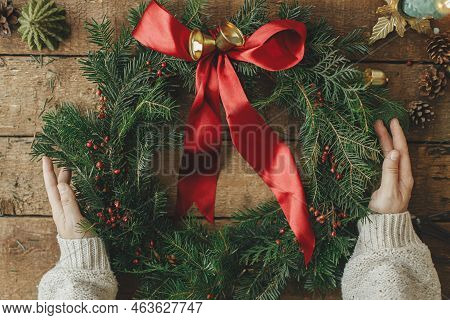 Woman Holding Stylish Christmas Wreath With Red Ribbon, Golden Bells And Berries On Rustic Wooden Ta
