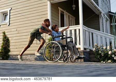Side View Portrait Of Black Teenage Boy Pushing Littlle Brother In Wheelchair While Having Fun Toget