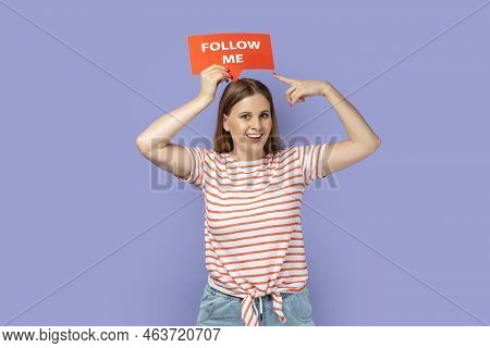 Portrait Of Delighted Smiling Blond Woman Holding Red Card With Follow Me Inscription Above Head, Po