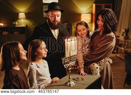 Portrait Of Modern Jewish Family Looking At Silver Menorah Candle During Hanukkah Celebration In Coz