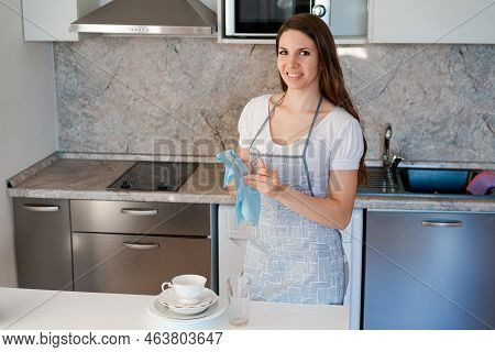 Wipe Dry. Lovely Woman Standing In Kitchen Wearing An Apron And Wiping A Dry Plate With A Kitchen To