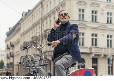 Middle-aged Man With Gray Hair Wearing Suit Riding A Rental Bike To Work Stopping To Make A Call. Ci