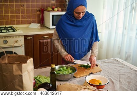 Pleasant Middle-eastern Muslim Woman Housewife, With Her Head Covered In A Blue Hijab, Chopping Gree