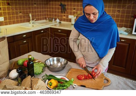 Charming Arab Muslim Housewife, Wearing A Blue Hijab, Stands At The Kitchen Table With A Lot Of Orga