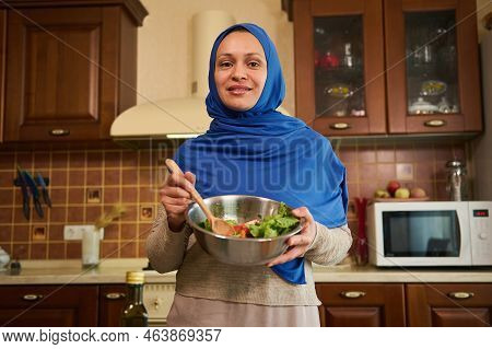 Charming Muslim Arab Young Woman With Head Covered In Blue Hijab, Standing In The Home Kitchen With 