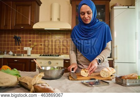 Muslim Arab Housewife With Head Covered In A Blue Hijab, Cutting Loaf Of Whole Grain Wheat Bread, Ma