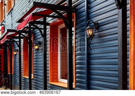 Wooden Facade Of Building With Retro Lanterns Illuminating The House With Windows And Red Visor, The