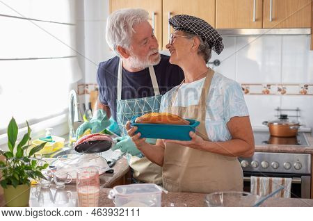 Lovely Senior Couple Kissing In The Home Kitchen While Wife Holds A Freshly Baked Homemade Plumcake 