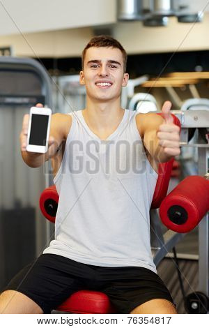 sport, bodybuilding, lifestyle, technology and people concept - smiling young man showing smartphone and thumbs up gesture in gym