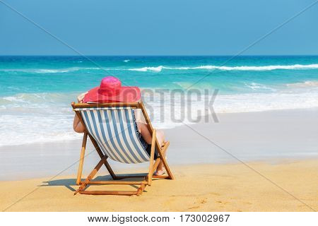 Happy woman in red sunhat on the beach sitting on deckchair and looking into the sea.Vacation and travel concept.