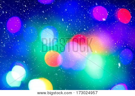 Defocused colorful ligths of Christmas tree. Multicolored bokeh lights during a snowfall. Background for greeting card. Winter composition. Happy holiday mood. Xmas and New Year fairy tale wallpaper.