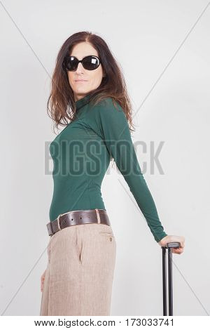 portrait of brown hair adult woman with green sweater beige trousers and sunglasses looking holding suitcase handle isolated on white